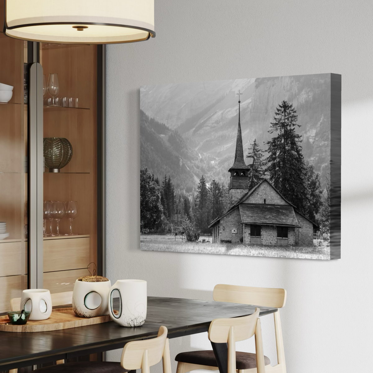 A black and white canvas wall art print depicting a mountain village church, displayed on a light gray wall in a modern dining room setting.