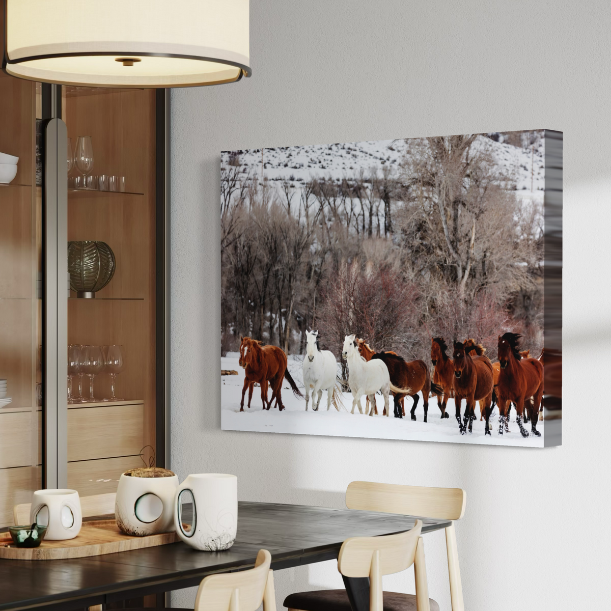 A canvas wall art print showing a herd of horses in a snowy outdoor scene, displayed above a dining table in a modern interior with chairs, mugs, and shelving visible.