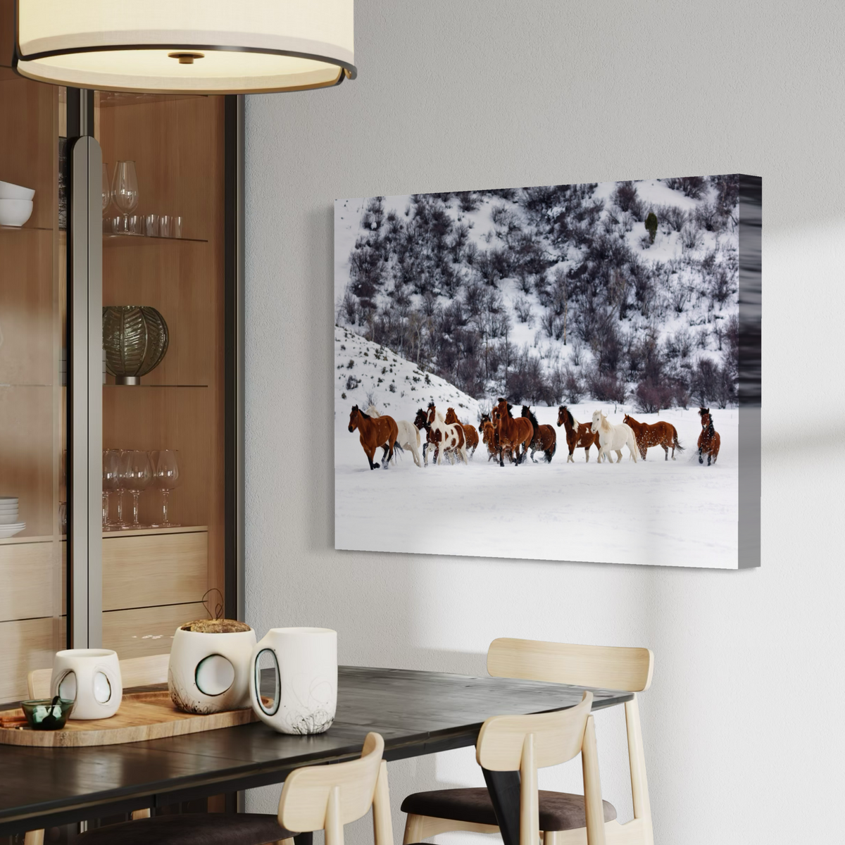 A canvas wall art print showing a herd of horses running through a snowy landscape, displayed on a light gray wall above a dining table in a modern interior setting.