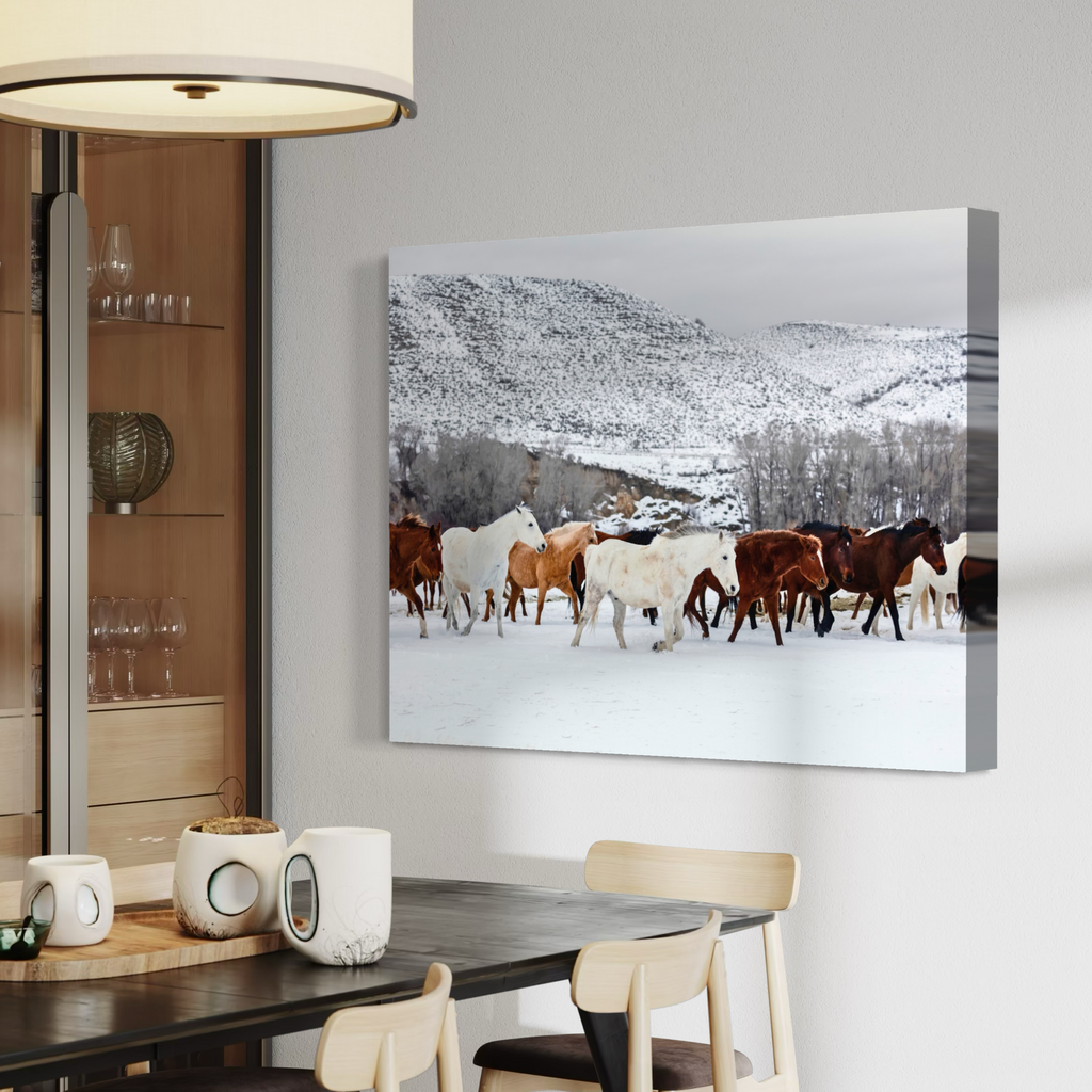 A canvas wall art print depicting a herd of cows and horses walking through a snowy mountain landscape, displayed on a light gray wall above a dining table in a modern kitchen setting.