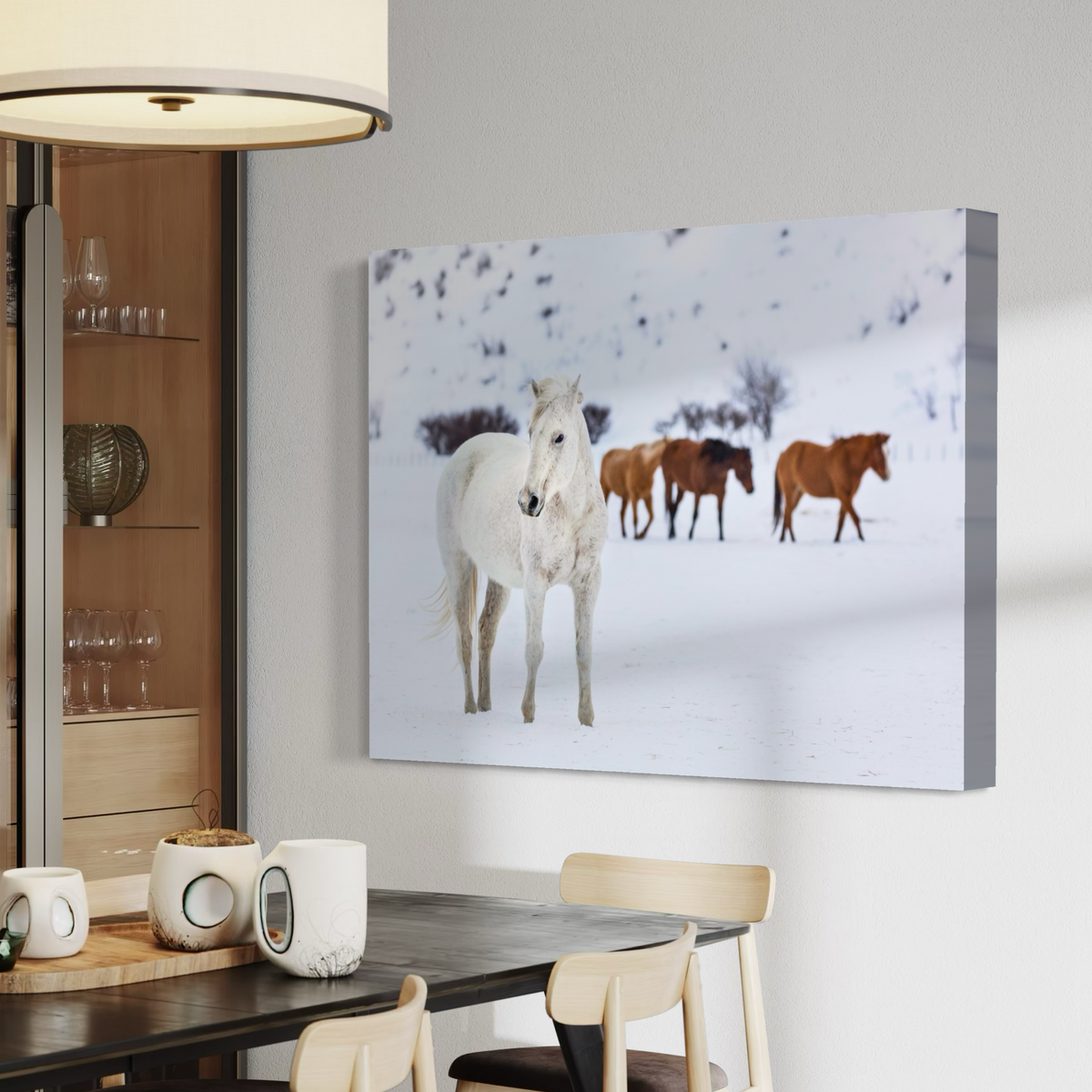 A canvas wall art print showing a snowy winter scene with a white horse in the foreground and two brown horses behind it, displayed above a dining table in a modern interior.