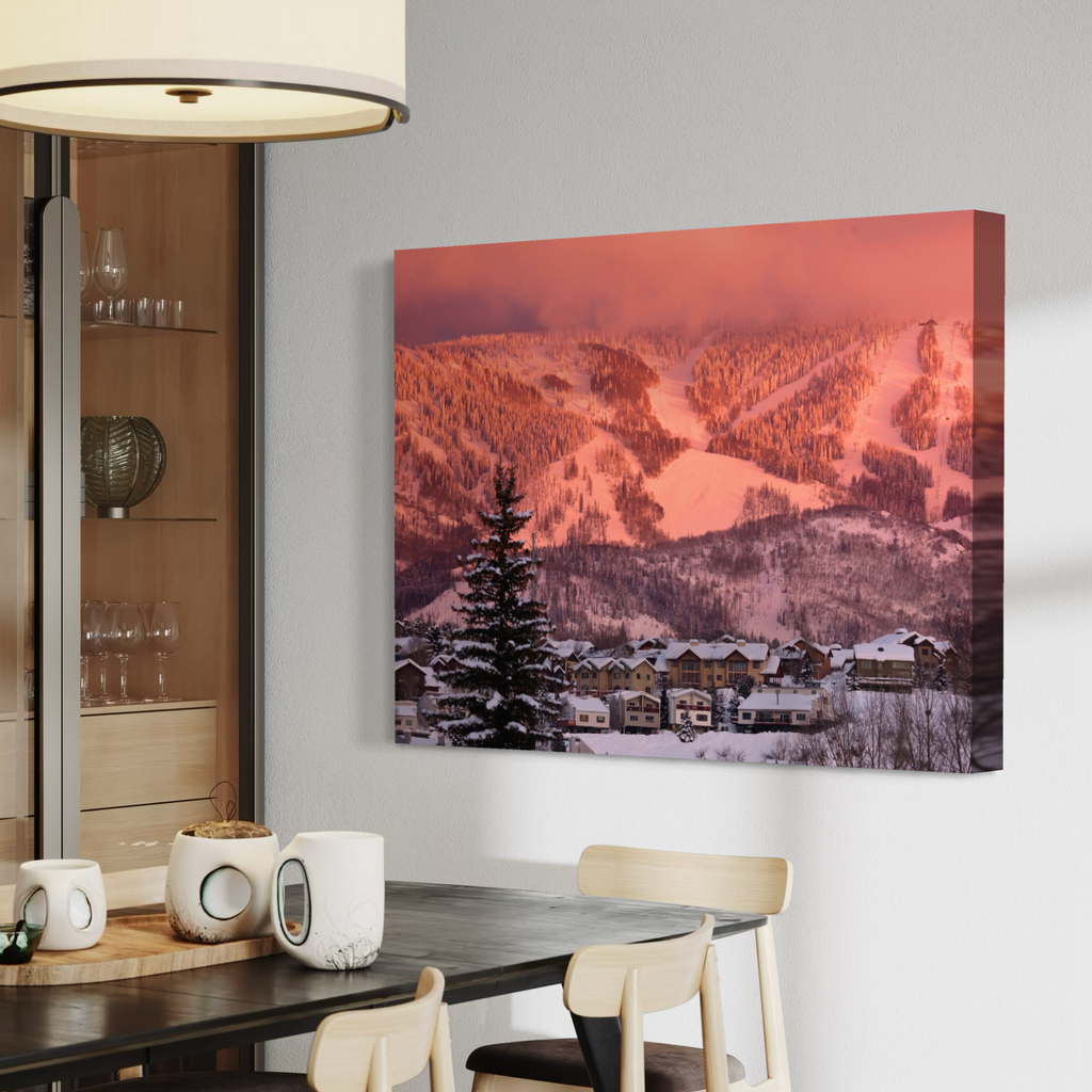 A canvas wall art print depicting a snowy mountain village under a pink-orange sky, displayed on a light gray wall in a modern dining room setting.