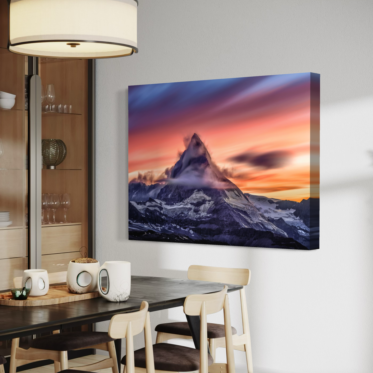 A rectangular canvas wall art print showing a mountain peak at sunset hangs on a light gray wall above a dining table in a modern interior. The artwork features a dramatic sky with purple, pink, orange, and blue tones over a snow-dusted mountain.