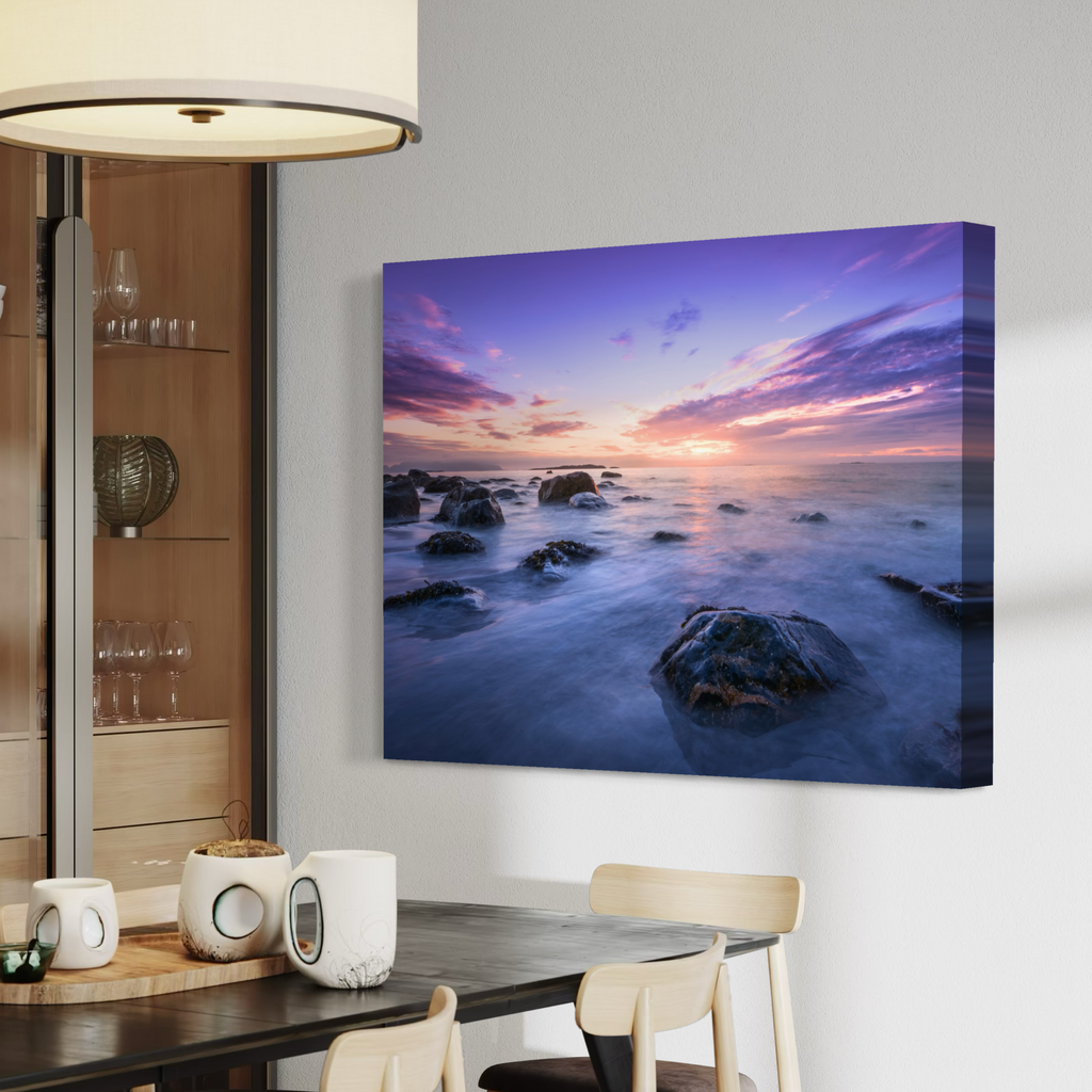 A canvas wall art print showing a rocky ocean sunset in purple, pink, and blue tones, displayed on a light gray wall above a dining table in a modern interior setting.