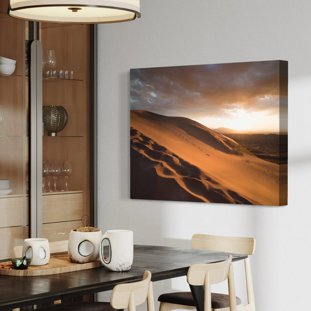 A rectangular canvas wall art print showing a desert landscape at sunset hangs on a light gray wall above a dining table in a modern interior setting.