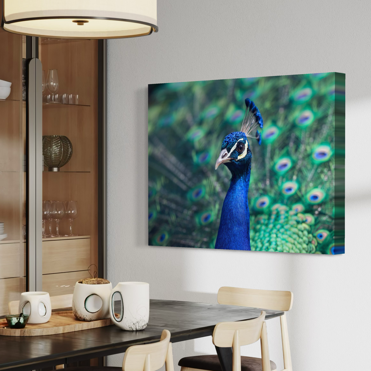 A canvas wall art print depicting a close-up peacock with vivid blue neck and green peacock-feather patterns in the background, displayed on a light gray wall above a dining table in a modern interior setting.