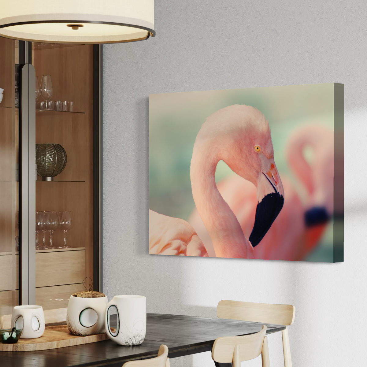 A canvas wall art print depicting a close-up pink flamingo, displayed on a light gray wall in a modern dining room setting with a table, chairs, and a lamp visible.