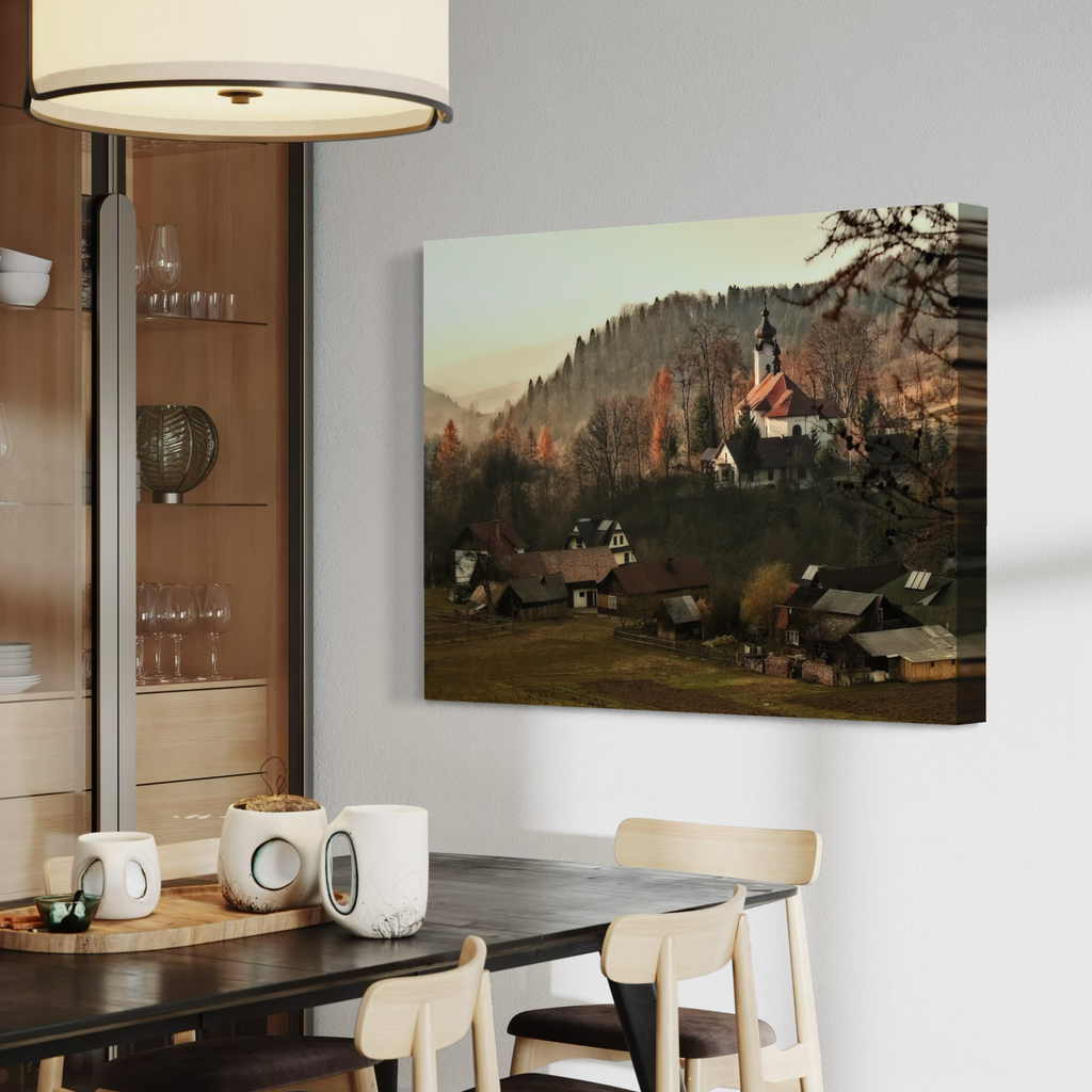 A rectangular wall art print showing a mountain village with a church, trees, and houses, displayed on a light gray wall above a dining table in a modern interior setting.