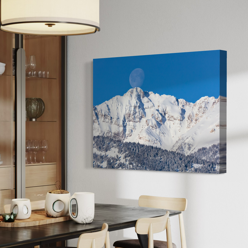A canvas wall art print depicting a snowy mountain peak under a bright blue sky with a full moon, displayed on a light gray wall in a modern dining room setting.