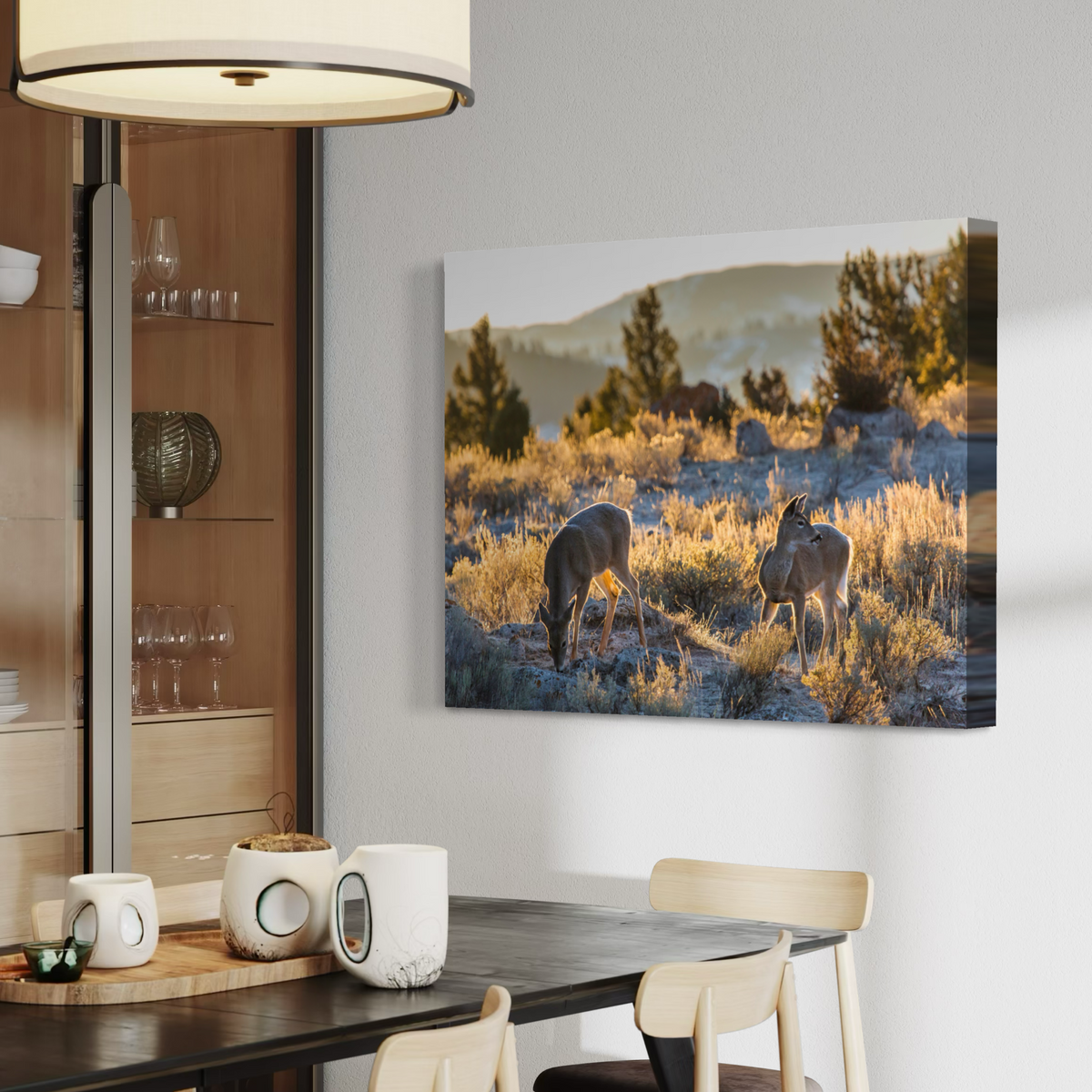 A rectangular canvas wall art print showing two deer in a sunlit grassy landscape hangs on a light gray wall above a dining table in a modern interior setting.
