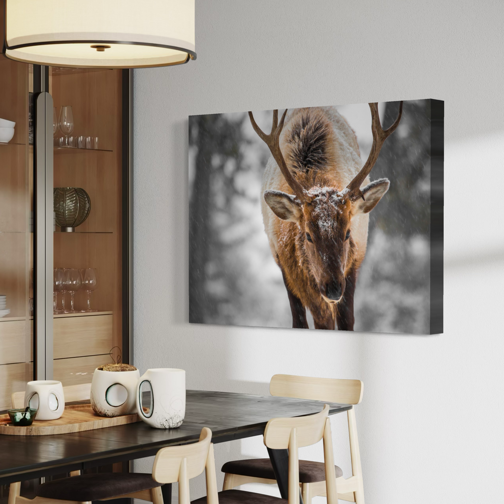 A large rectangular canvas wall art print showing a close-up of a deer with antlers in a snowy, monochrome-toned winter scene, displayed above a dining table in a modern interior.