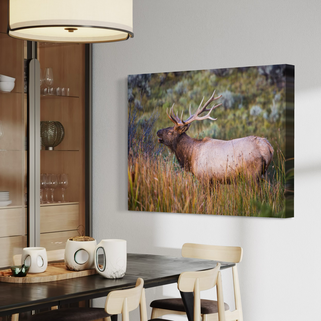 A rectangular canvas wall art print showing a large elk standing in tall grass, displayed on a light gray wall above a dining table in a modern interior setting.