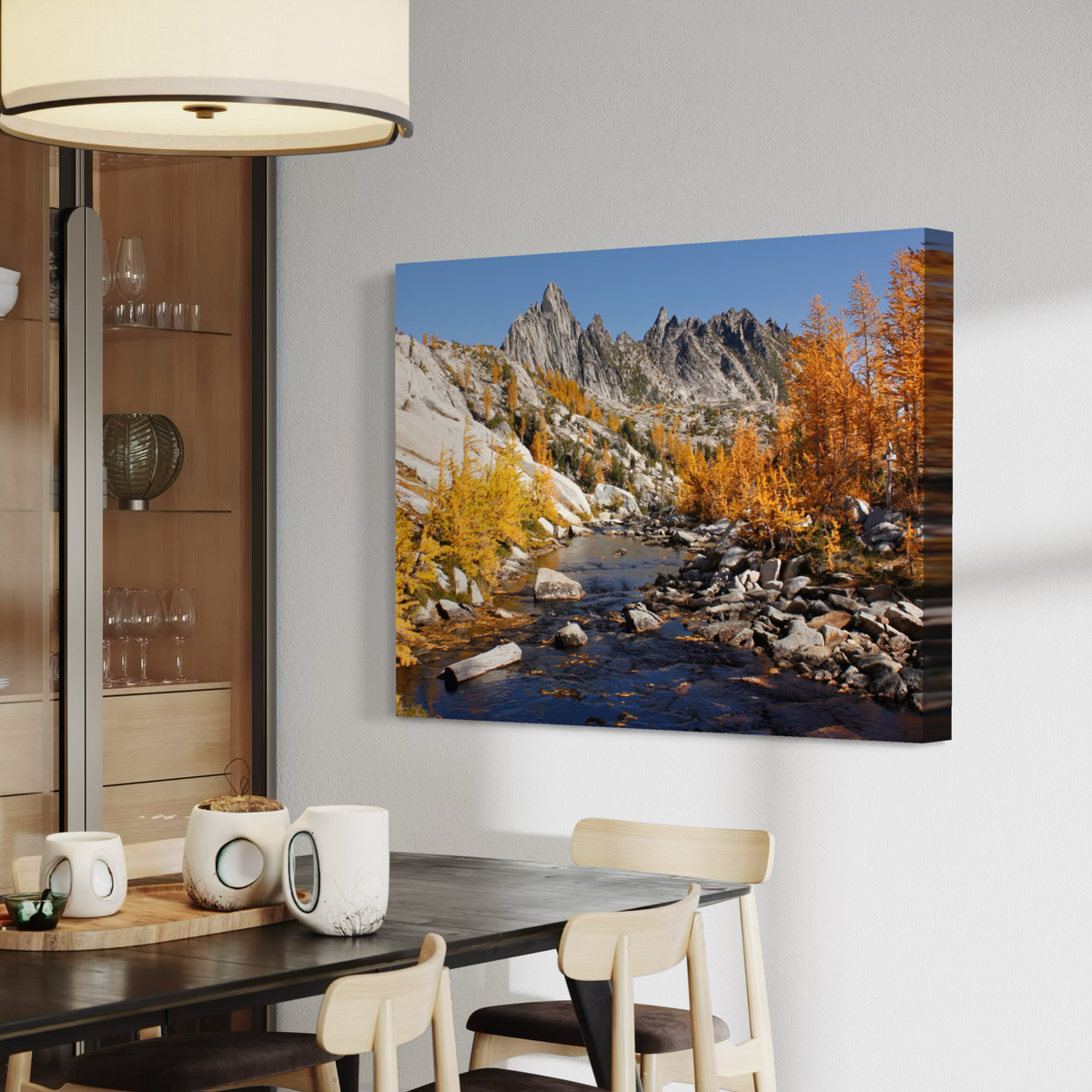 A canvas wall art print showing a mountain landscape with a rocky stream, blue sky, and golden autumn trees, displayed above a dining table in a modern interior.