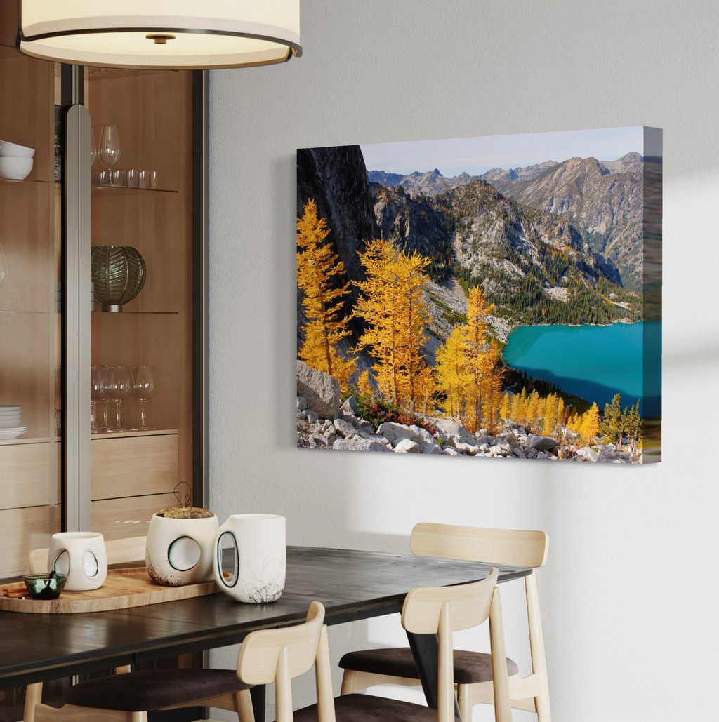 A rectangular wall art canvas print showing a mountain landscape with bright yellow trees and a turquoise lake, displayed above a dining table in a modern interior setting.