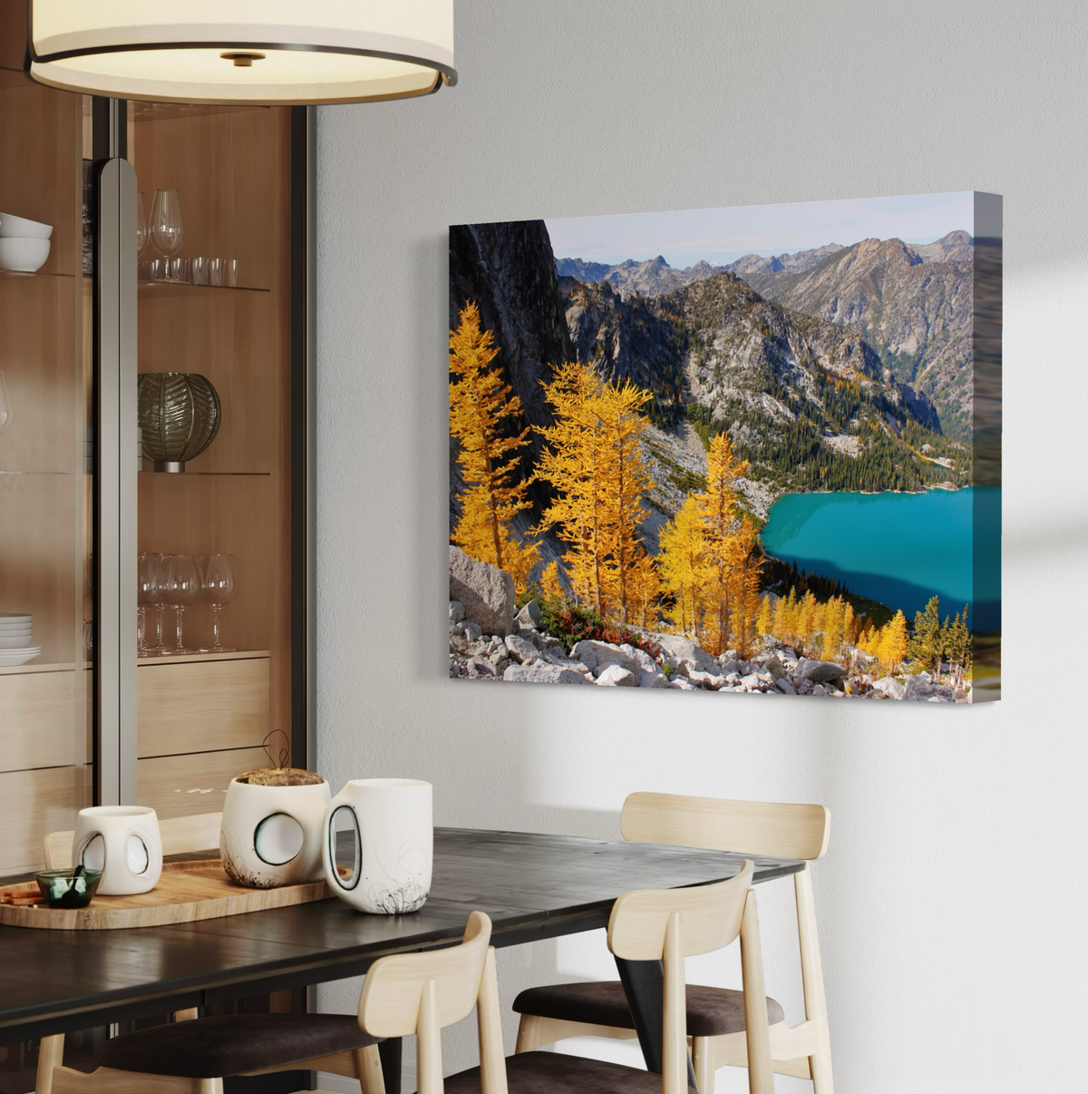 A rectangular wall art canvas print showing a mountain landscape with bright yellow trees and a turquoise lake, displayed above a dining table in a modern interior setting.