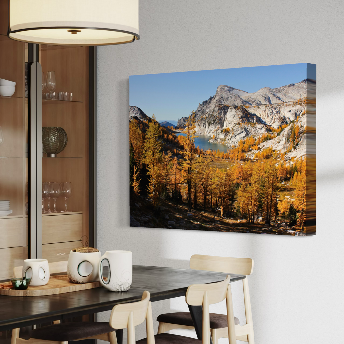A canvas wall art print showing a mountain landscape with golden autumn trees and a blue lake, displayed above a dining table in a modern interior room.