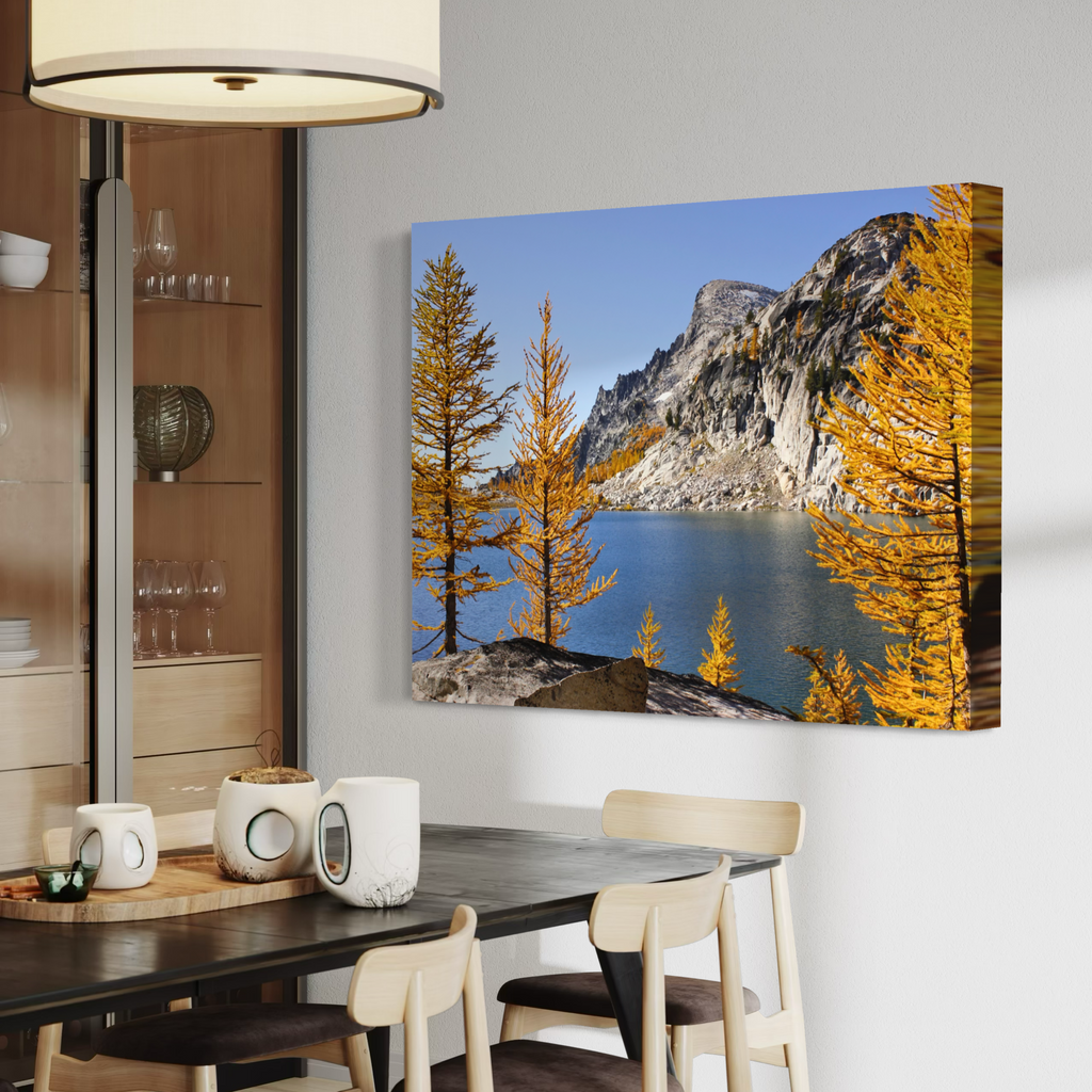 A canvas wall art print showing a mountain lake scene with golden yellow trees under a blue sky, displayed on a light gray wall above a dining table in a modern interior.