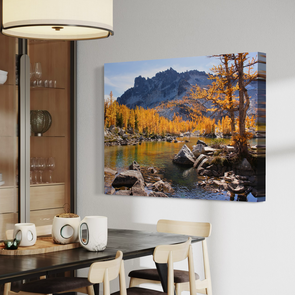 A wall art canvas showing a mountain lake landscape with golden autumn trees, displayed above a dining table in a modern interior room.