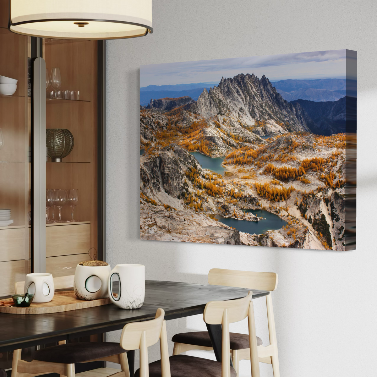 A canvas wall art print showing a mountain landscape with rocky peaks, blue lakes, and golden autumn trees, displayed above a dining table in a modern interior.