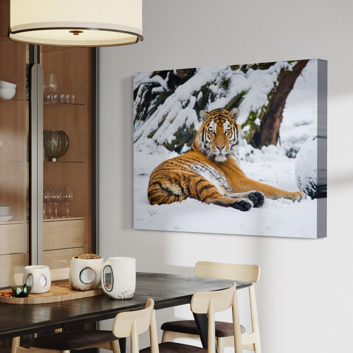 A wall art canvas print showing a tiger lying in a snowy outdoor setting, displayed above a dining table in a modern interior room.