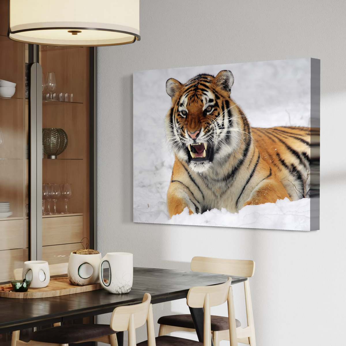A rectangular canvas wall art print showing a close-up tiger in white snow, displayed on a light gray wall above a dining table in a modern interior setting.