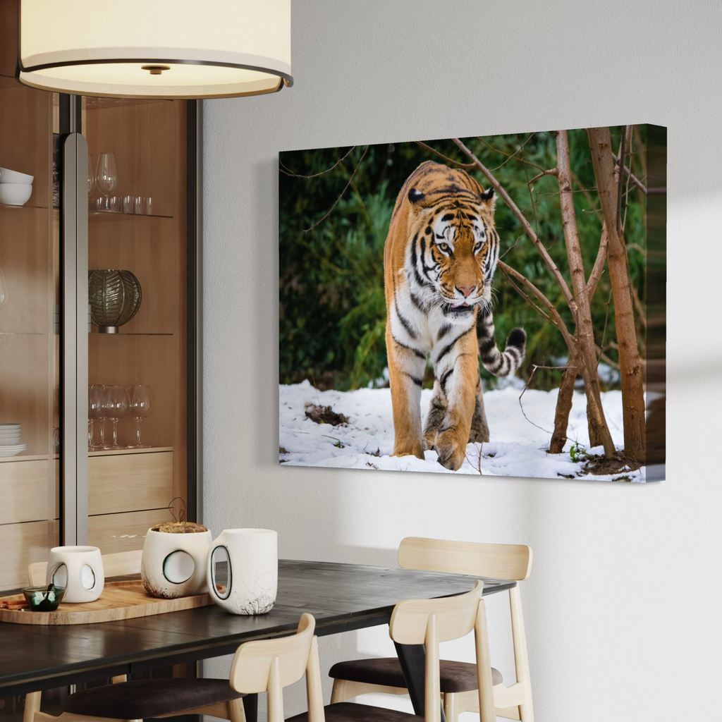 A wall art canvas print showing a tiger walking through snow with trees in the background, displayed above a dining table in a modern interior room.