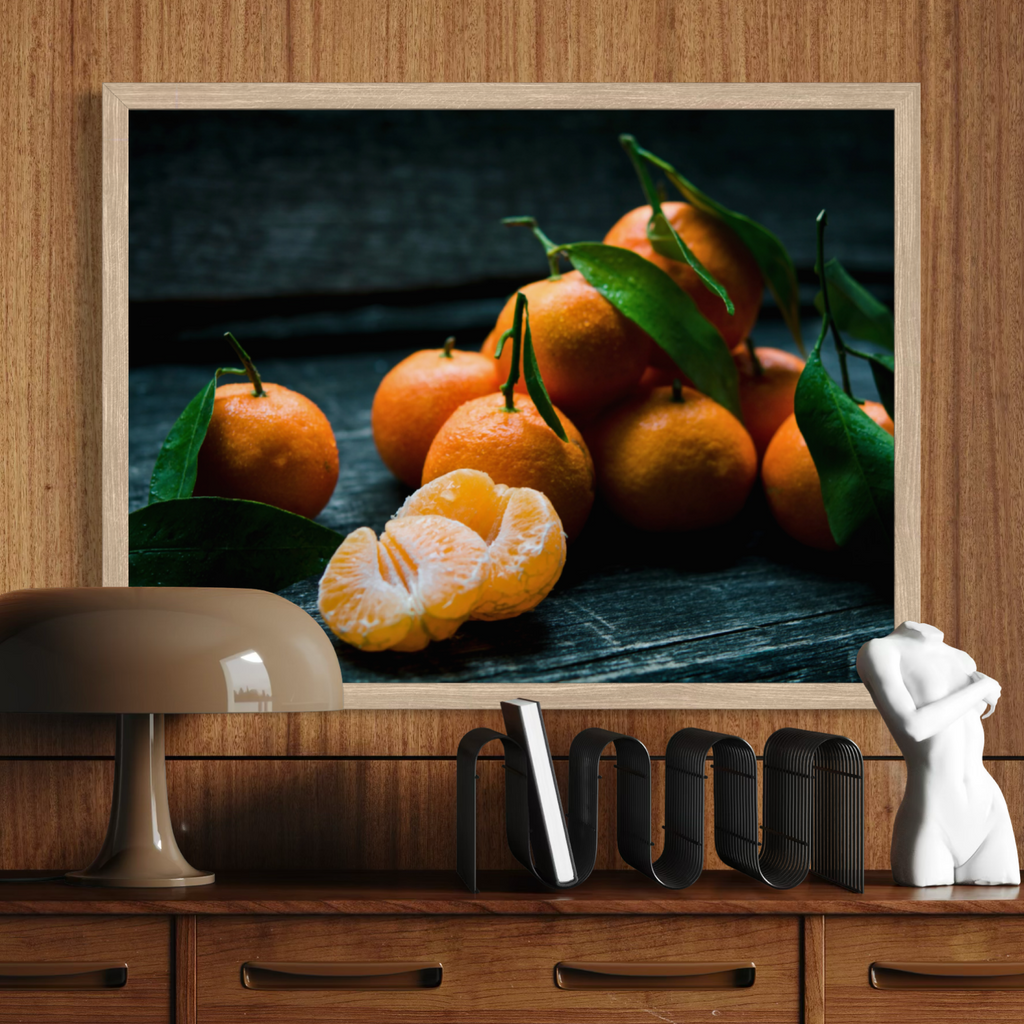 A framed wall art print showing a close-up still life of orange clementines with green leaves, displayed on a wooden shelf above a lamp and decorative objects.