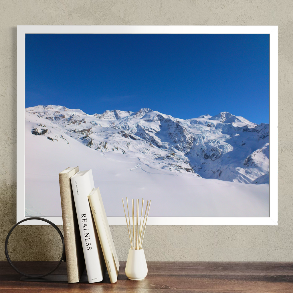 A large framed wall art print showing a snowy mountain landscape under a vivid blue sky, displayed on a beige wall above a wooden surface with books and a small vase.