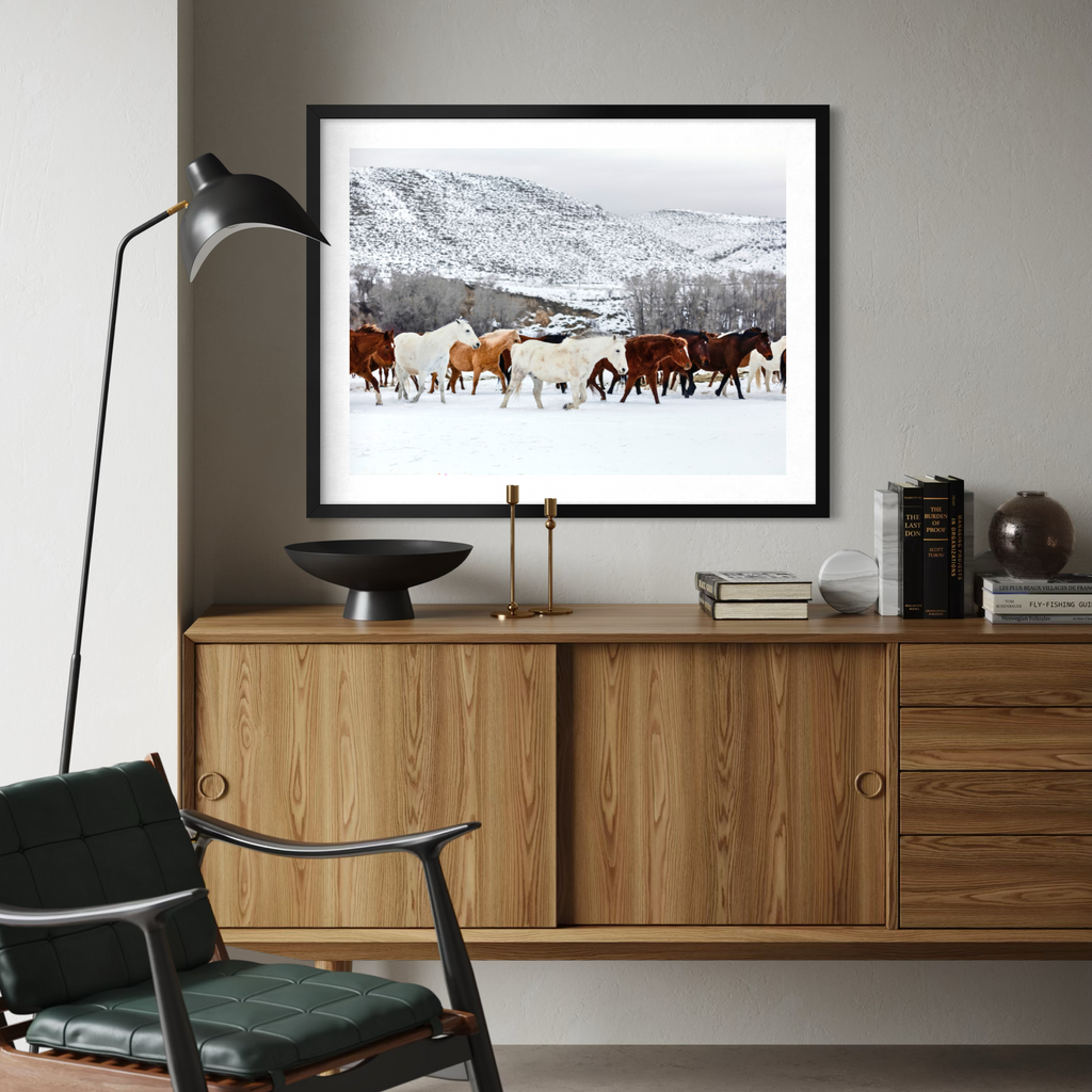 A framed art print depicting a herd of horses walking through a snowy mountain landscape, displayed on a wooden sideboard in a modern living room setting.