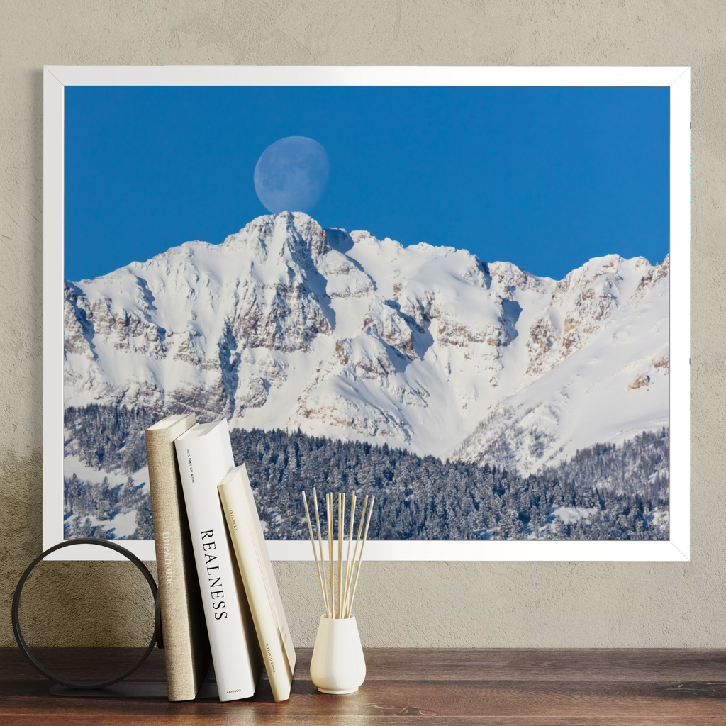 A framed wall art print showing a snowy mountain range under a bright blue sky with a visible moon, displayed above a wooden surface with books and a small vase.