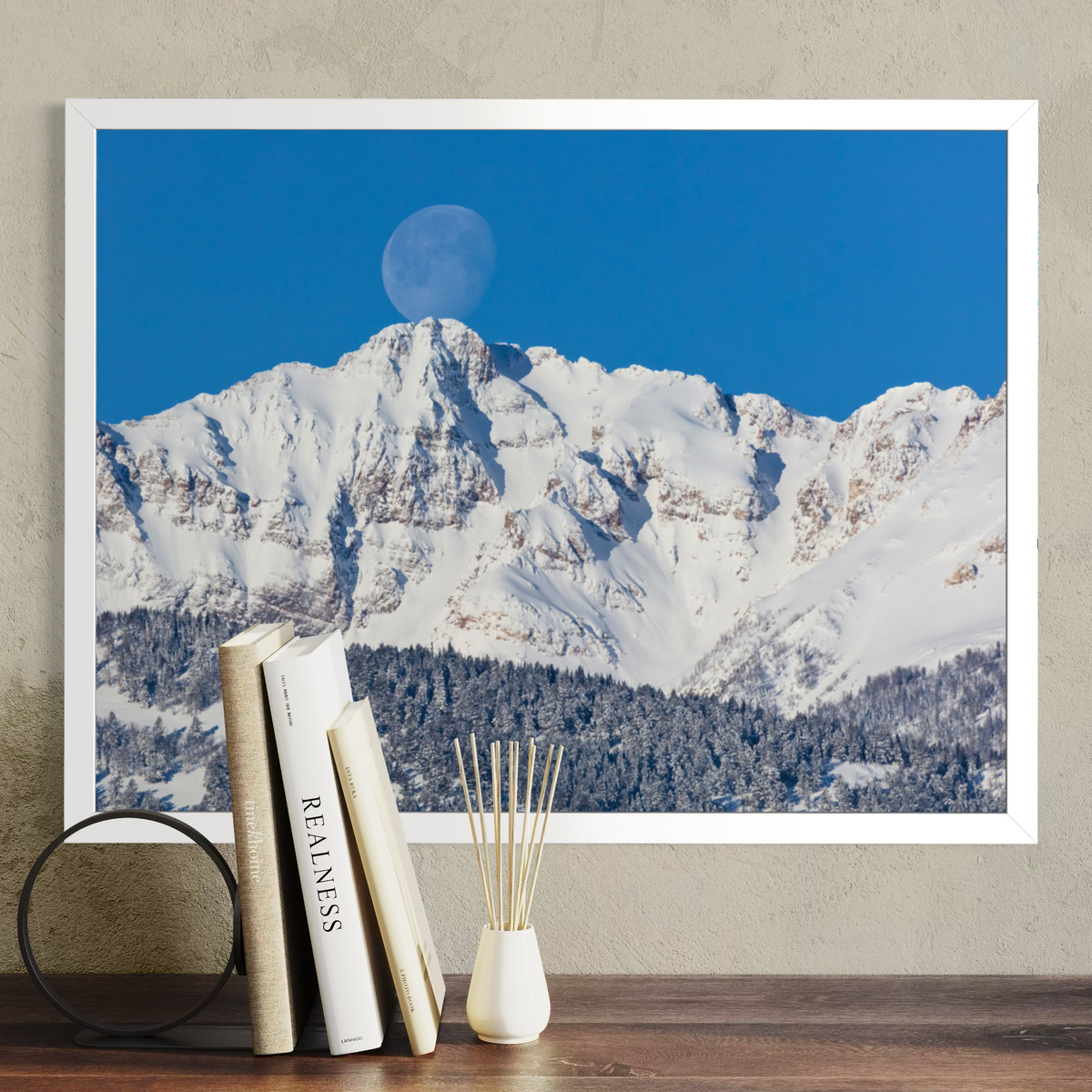 A framed wall art print showing a snowy mountain range under a bright blue sky with a visible moon, displayed above a wooden surface with books and a small vase.