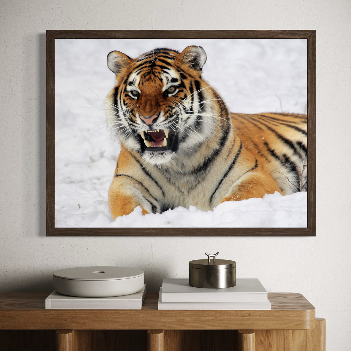 A dark wooden framed wall art print showing a close-up tiger in the snow, hung on a light wall above a wooden console table with books and decorative objects.