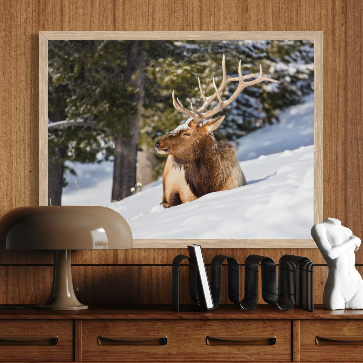 A wooden framed wall art print depicting a brown elk with large antlers sitting in a snowy forest, displayed on a wooden console table above a lamp and decorative objects.
