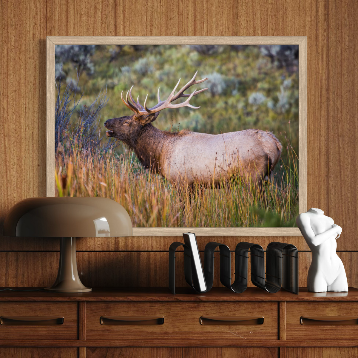 A wooden framed art print depicting a bull elk in a grassy field, displayed on a wooden wall above a matching wooden console table with a lamp, bookends, and a white figurine.