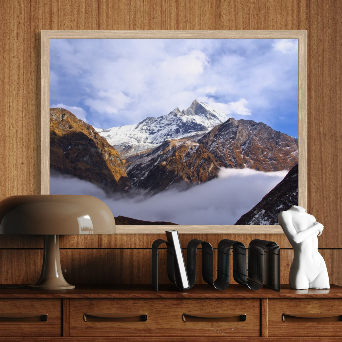 A framed wall art print depicting a snow-capped mountain peak above clouds, displayed on a wooden dresser against a wood-paneled wall. Decorative objects including a lamp, books, and a small white figurine are visible in the room setting.