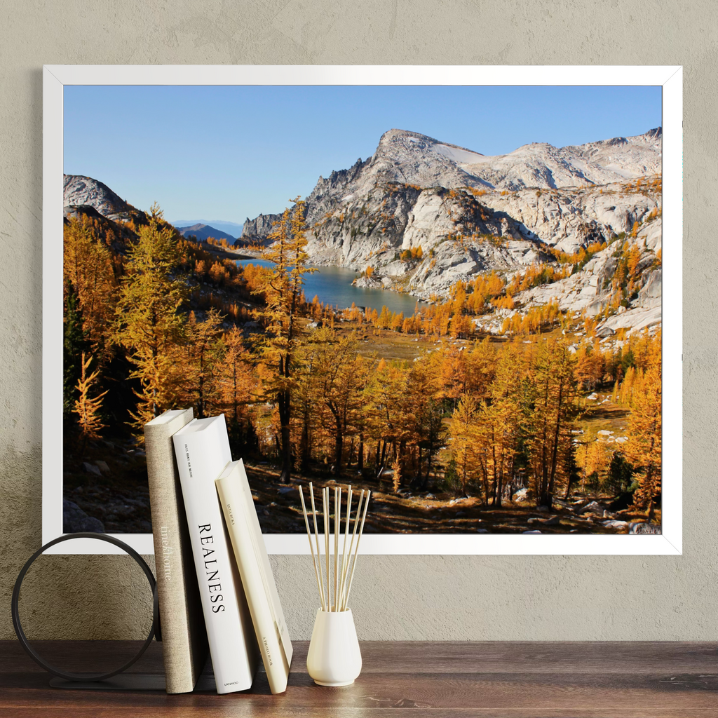A white framed landscape art print of a mountain valley with autumn trees and a lake, displayed on a wall above a wooden surface with books and a reed diffuser.