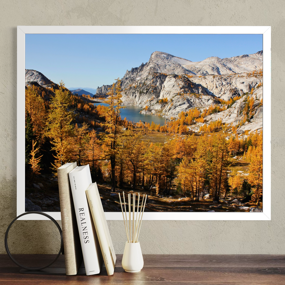 A white framed landscape art print of a mountain valley with autumn trees and a lake, displayed on a wall above a wooden surface with books and a reed diffuser.