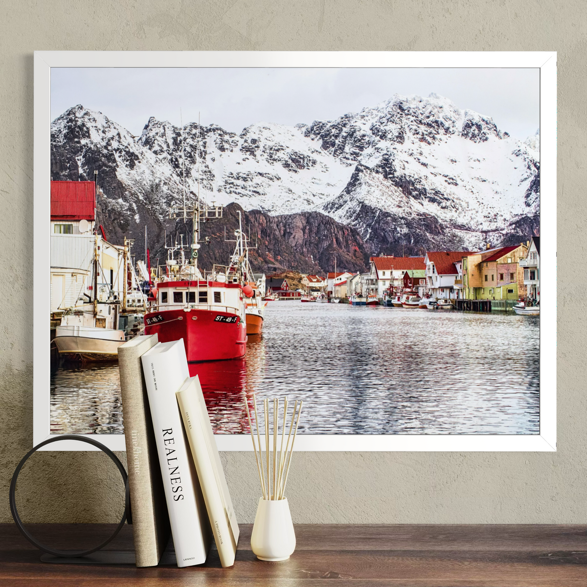 A framed art print depicting a scenic fjord with snow-capped mountains, boats, and colorful houses, displayed on a wall above a wooden surface with books and a vase.