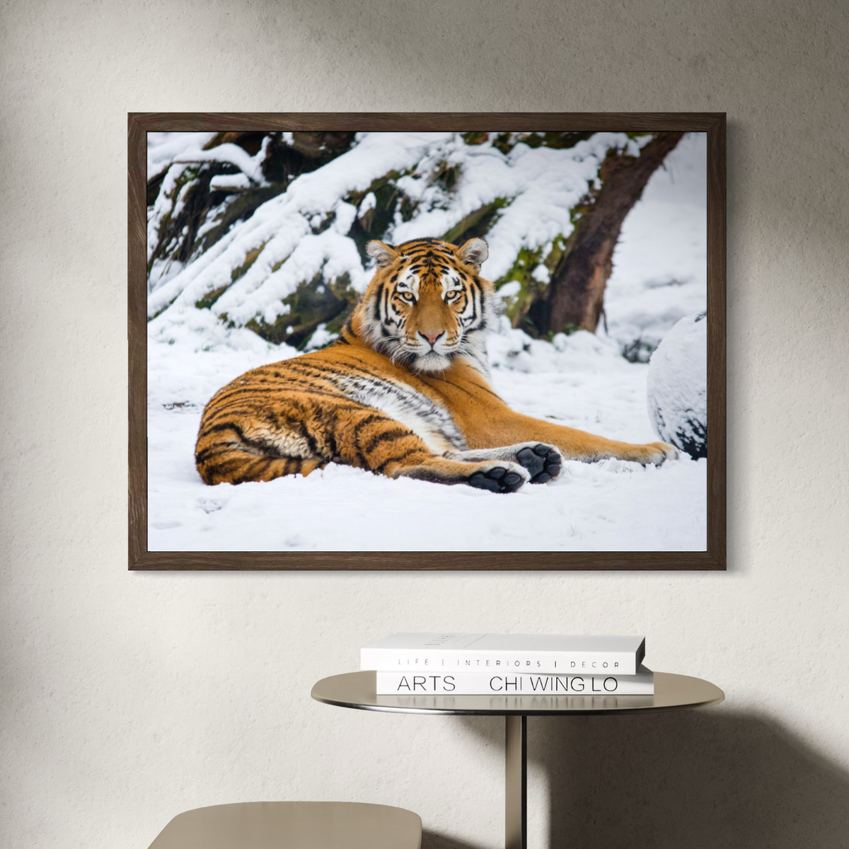 A wooden framed wall art print showing a tiger lying in a snowy outdoor scene, mounted on a light beige wall above a small round table with books.