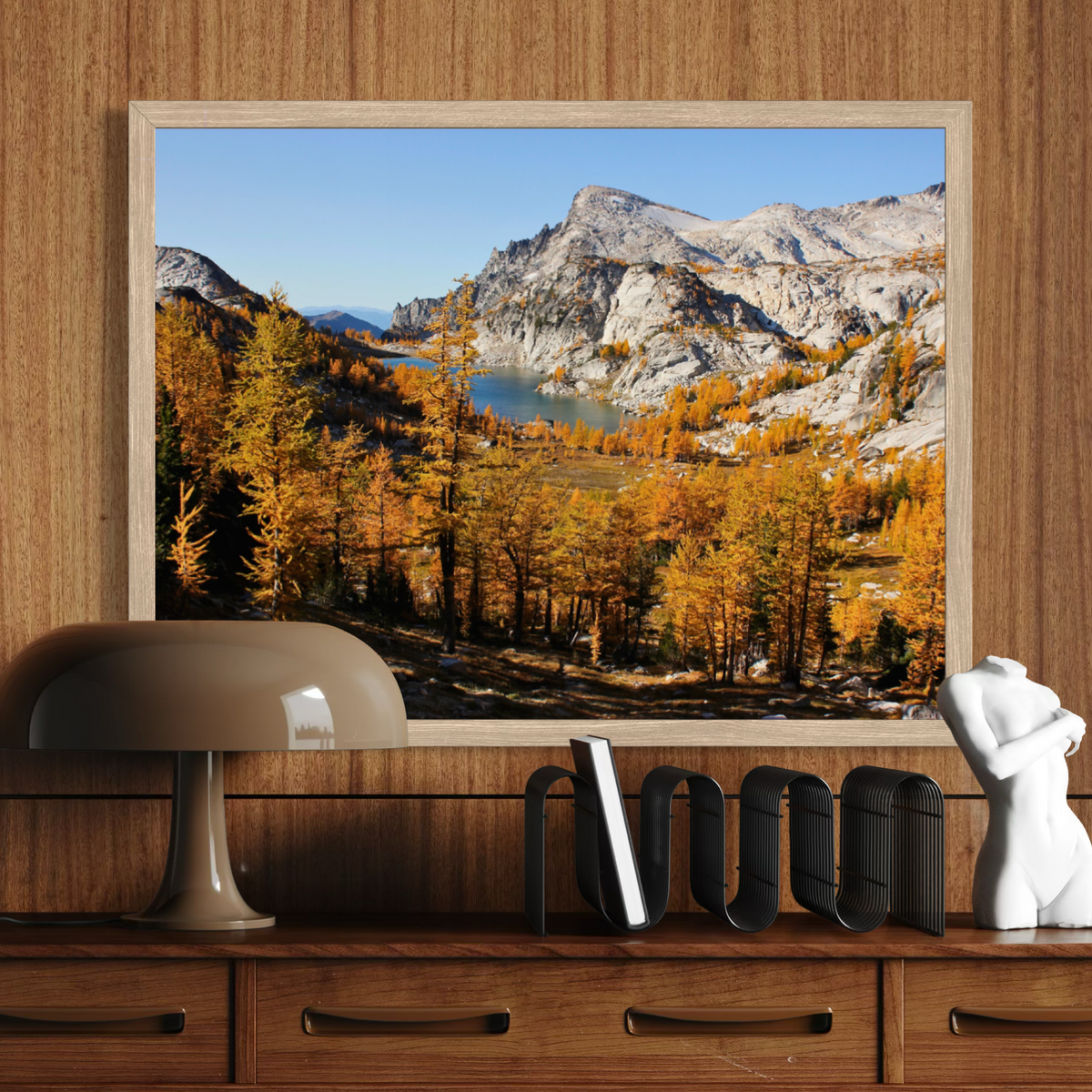 A wooden framed landscape art print depicting a mountain valley with golden autumn trees and a lake, displayed on a wooden console above a lamp and decorative objects.