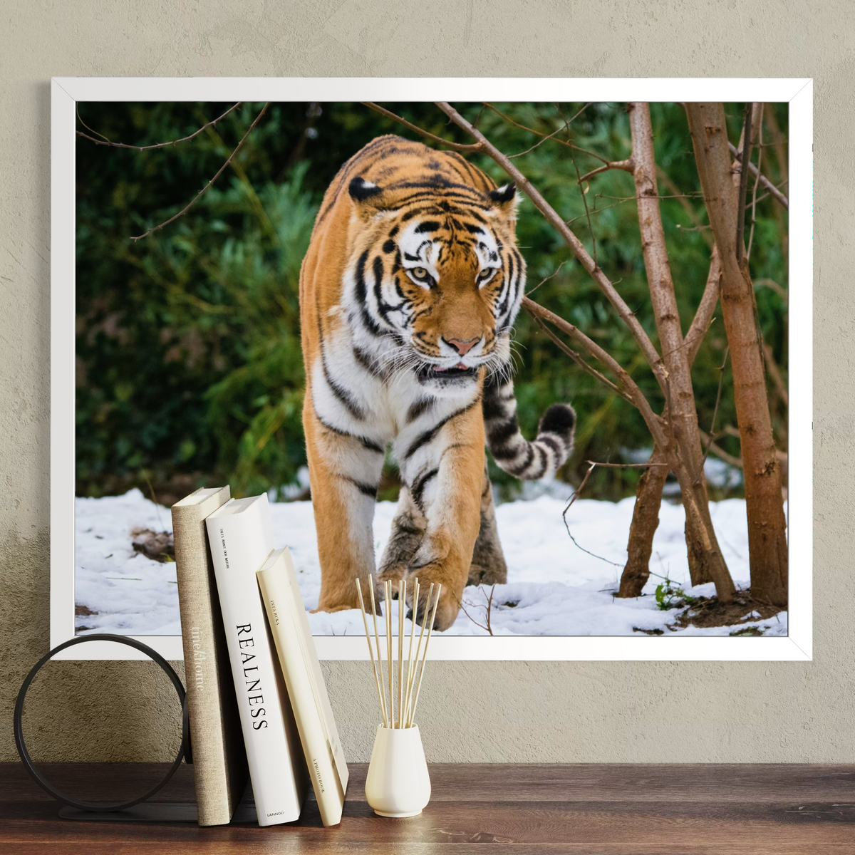 A framed wall art print showing a tiger walking through snowy trees, displayed on a beige wall above a wooden surface with books and a small vase.