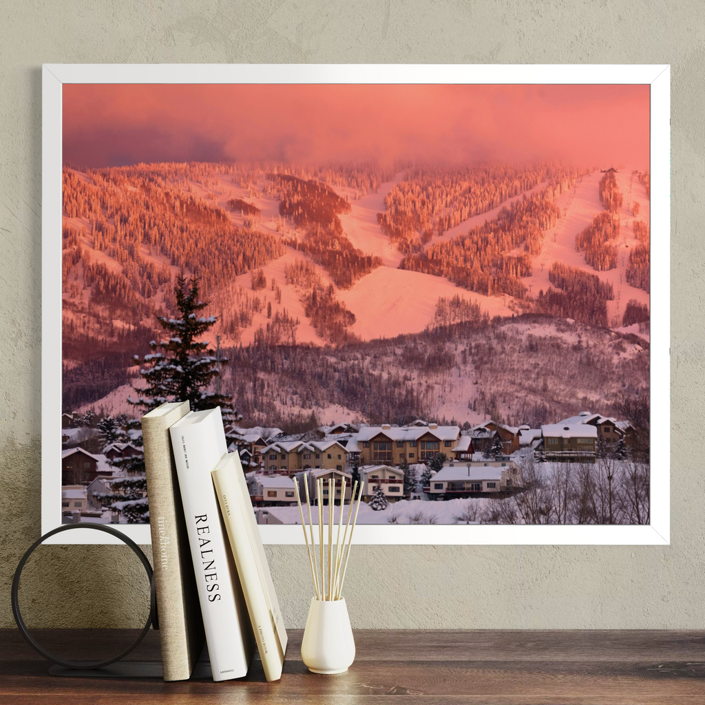A framed wall art print showing a snowy mountain landscape at sunset, displayed above a wooden surface with books and a small vase. The print is the product being sold, and the surrounding room decor is styled context.
