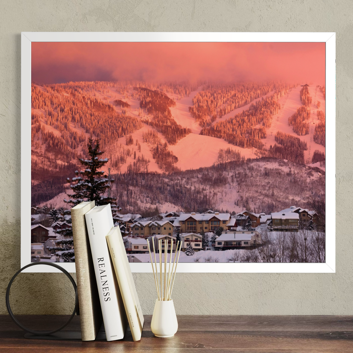 A framed wall art print showing a snowy mountain landscape at sunset, displayed above a wooden surface with books and a small vase. The print is the product being sold, and the surrounding room decor is styled context.