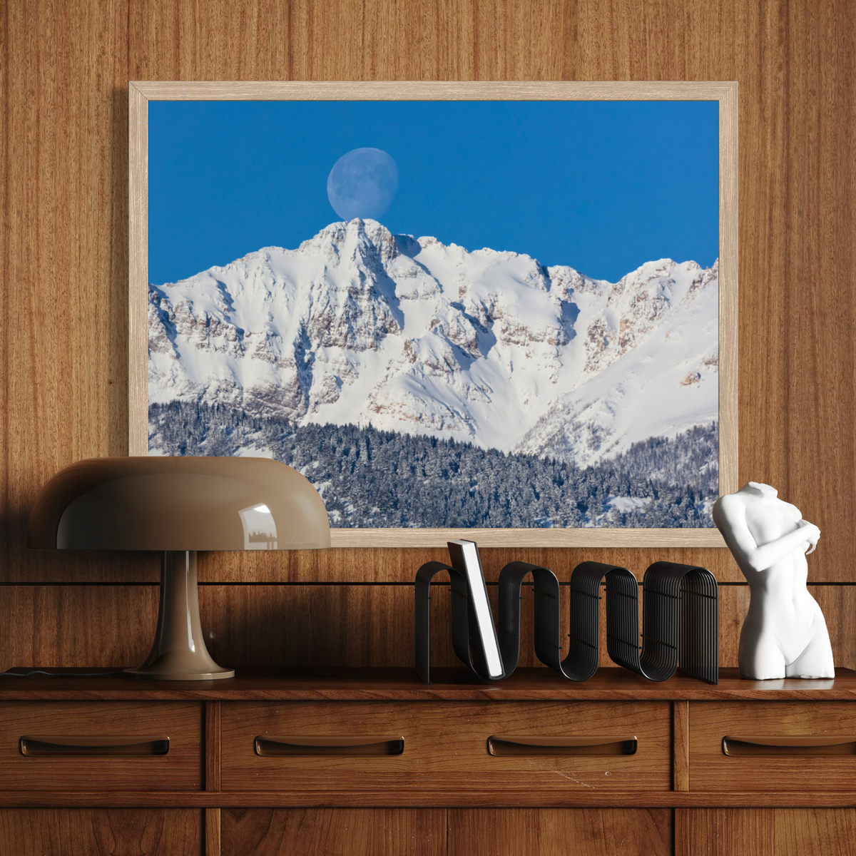 A wooden framed art print depicting a snow-covered mountain range under a clear blue sky with a visible moon, displayed on a wooden wall above a console table with a lamp, books, and a white sculpture.