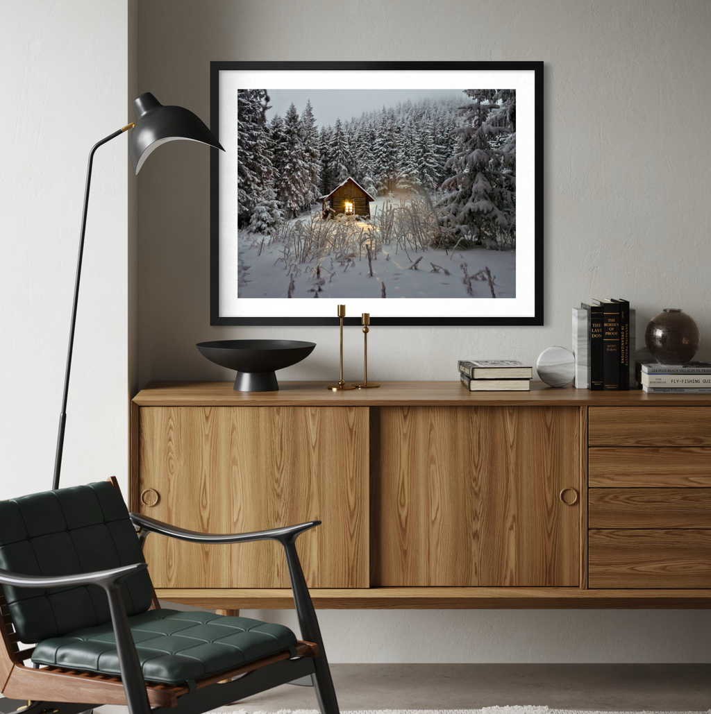 A framed art print depicting a cozy cabin in a snowy forest, displayed on a light gray wall above a wooden credenza in a modern living room setting.