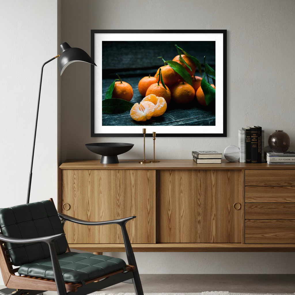 A framed wall art print depicting a still life of oranges with green leaves, displayed in a modern room setting above a wooden credenza.