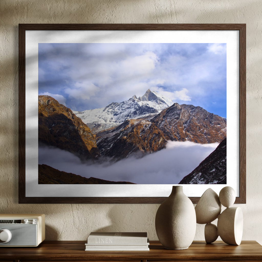 A framed mountain landscape print hangs on a textured beige wall above a wooden console table. The artwork shows snow-capped peaks, rocky brown slopes, and low clouds under a blue sky.