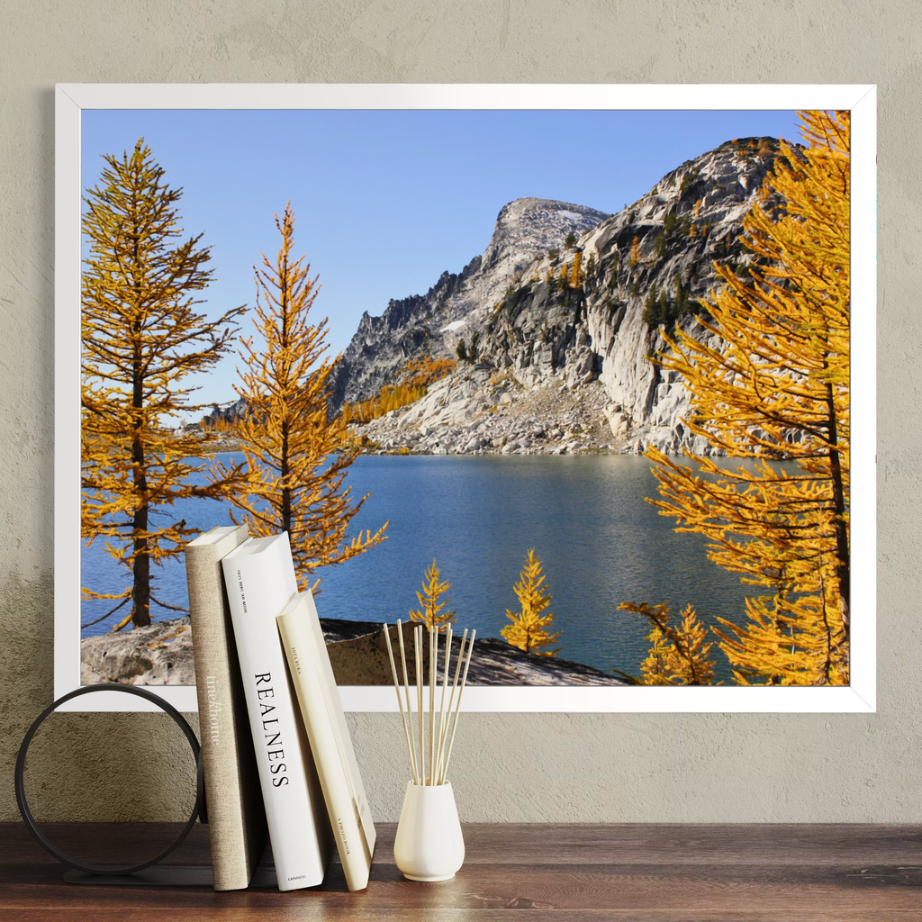 A white framed landscape art print depicting a mountain, blue lake, and golden-yellow autumn trees, displayed on a textured beige wall above a wooden table with books and a reed diffuser.