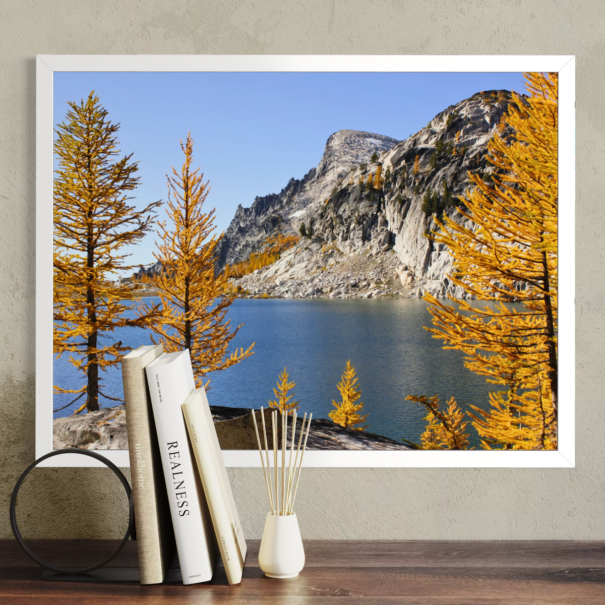 A white framed landscape art print depicting a mountain, blue lake, and golden-yellow autumn trees, displayed on a textured beige wall above a wooden table with books and a reed diffuser.