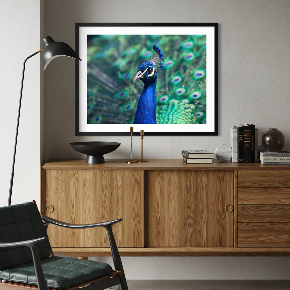 A framed wall art print of a peacock with green and blue feathers, displayed above a wooden console cabinet in a modern living room setting.